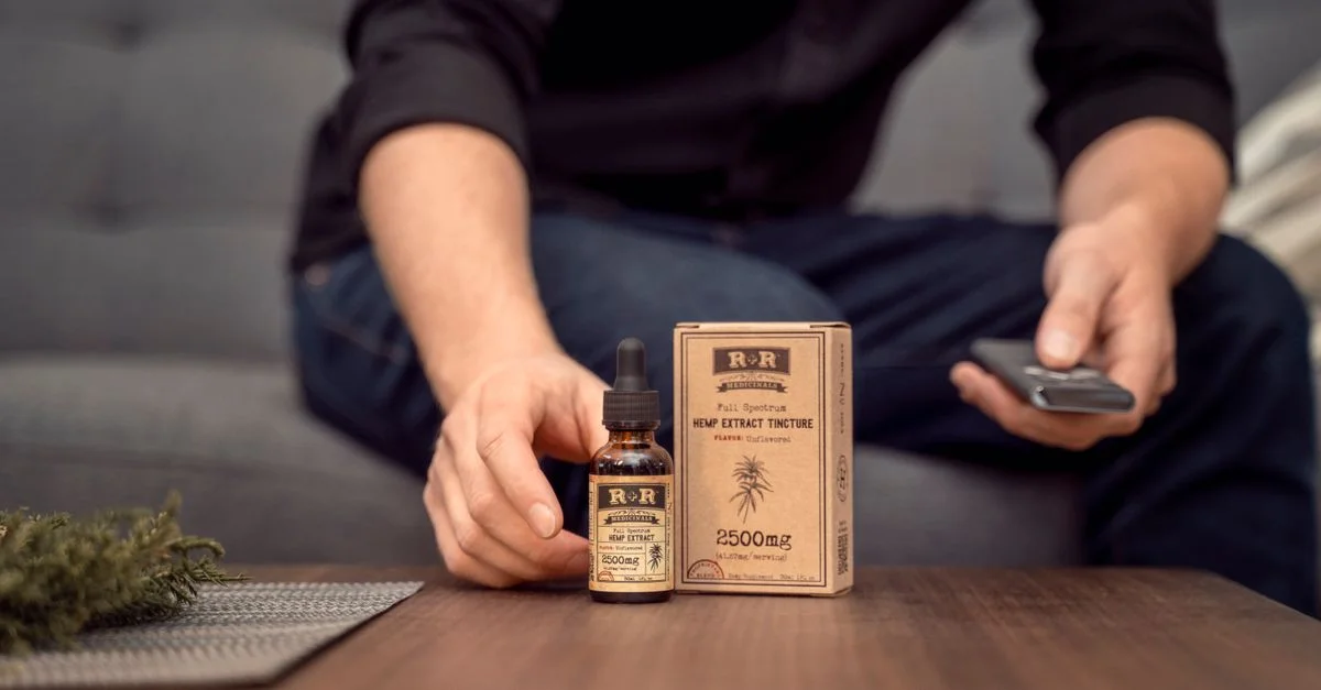 Homme tenant une bouteille d'huile de chanvre R&R à côté de l'emballage sur une table en bois dans le salon.