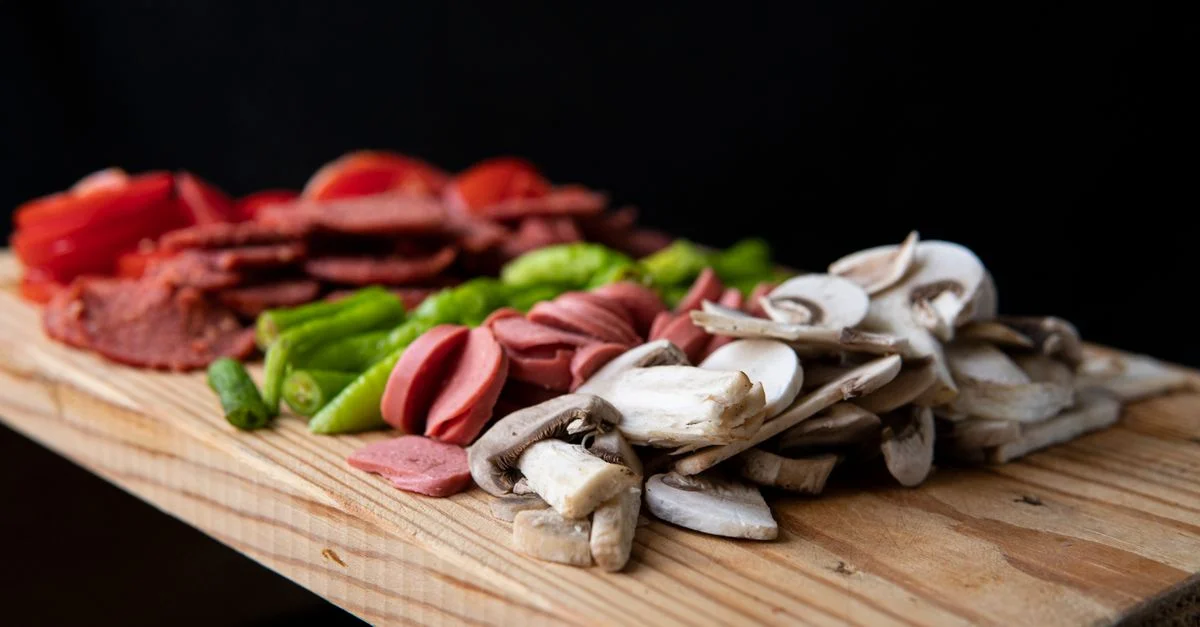 Une variété d'ingrédients frais incluant des champignons, des poivrons et des saucisses sur une planche à découper.
