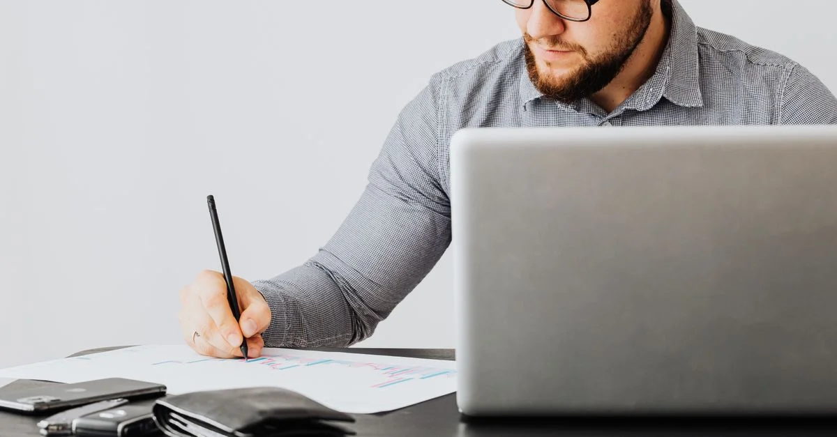 Un homme concentré analyse des documents financiers avec un ordinateur portable dans un bureau.