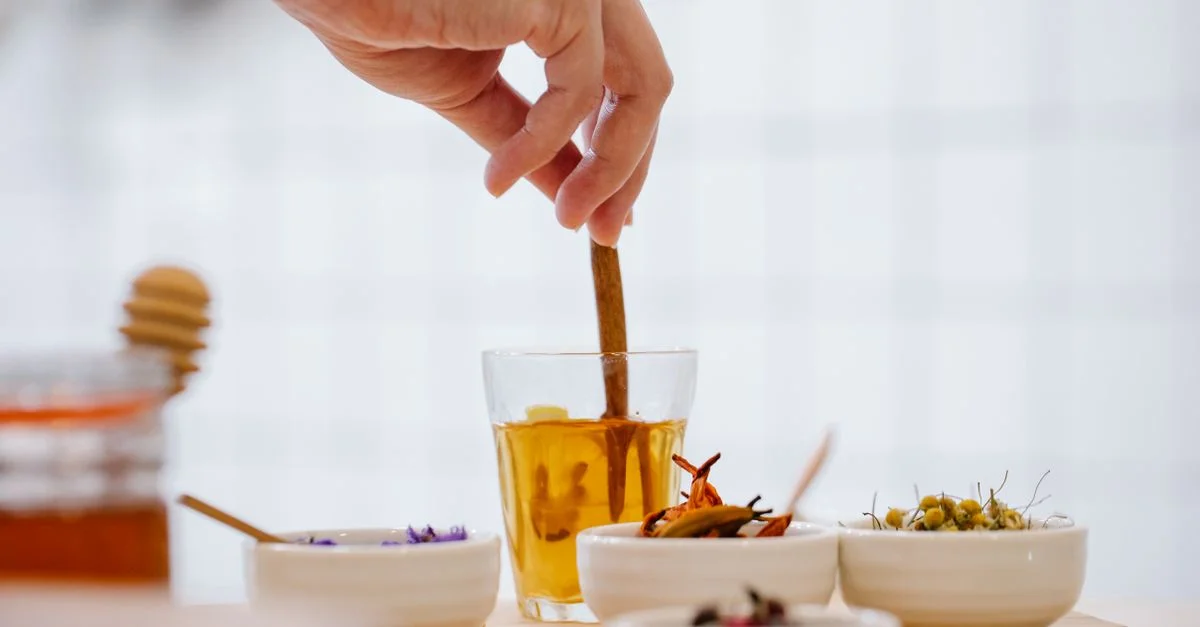 Une main remue du thé herbal dans un verre avec des bols d'épices et d'herbes sur un plan de travail de cuisine.