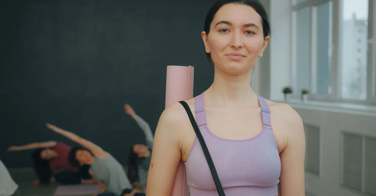 Jeune femme sûre d'elle, debout dans un studio de yoga, tenant un tapis, un groupe pratiquant en arrière-plan.
