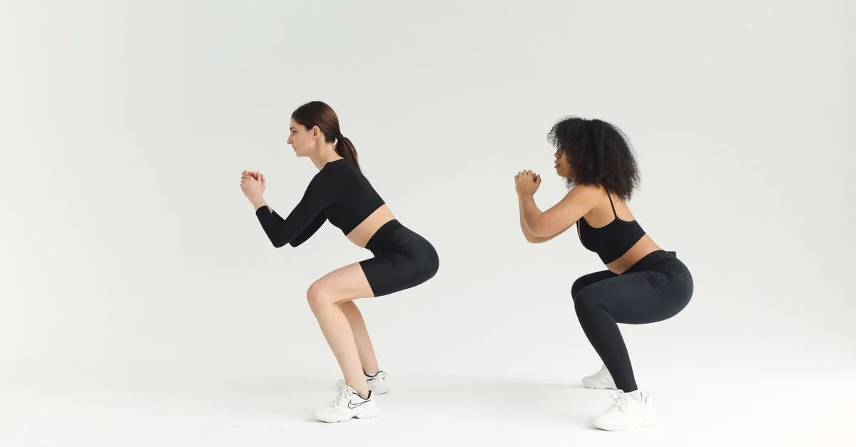 Deux femmes effectuant des squats en tenue de sport sur un fond uni, mettant en avant la forme physique et un mode de vie sain.